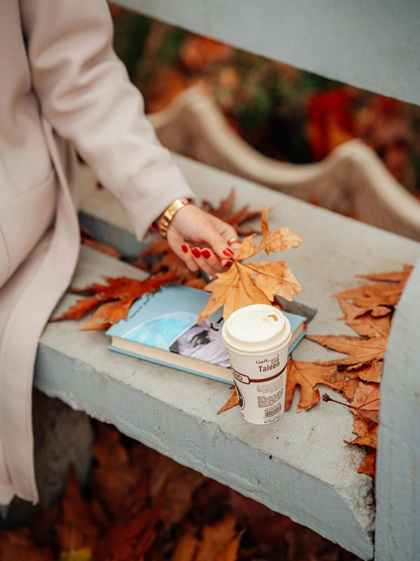 A fashionable girl in a stylish coat sits on a park bench surrounded by autumn leaves, with a book and a cup of coffee beside her.